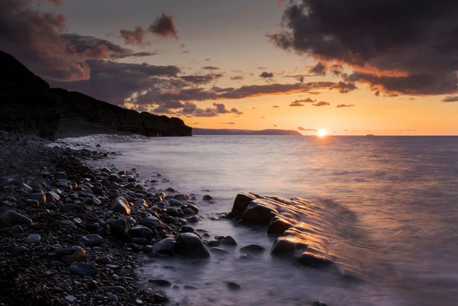 a sun sets on the horizon of a rocky beach